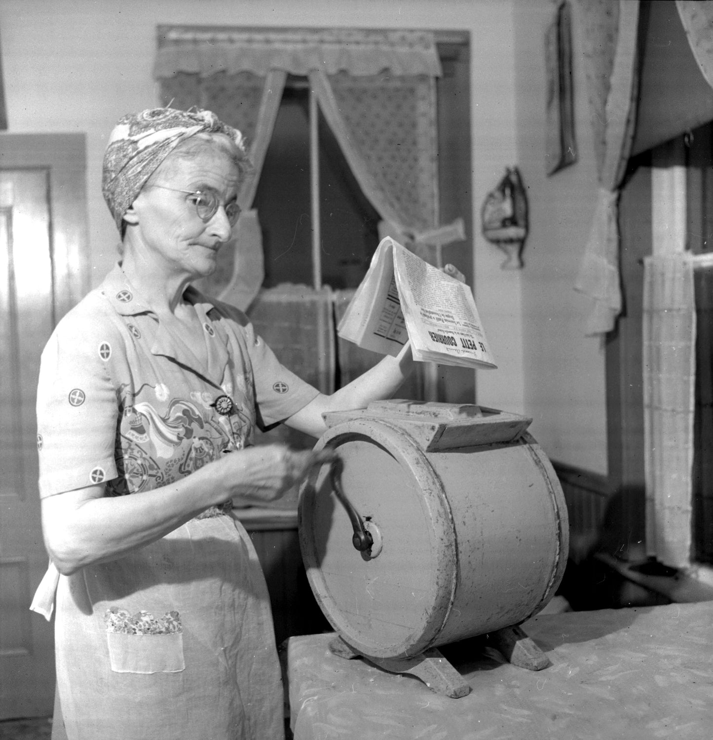 Mrs. Nicholas (Marie) Doucette churning butter while reading Le Petit Courier.