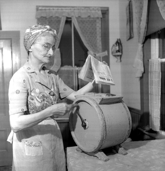 Mrs. Nicholas (Marie) Doucette churning butter while reading Le Petit Courier.